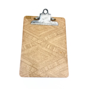 May include: A wooden clipboard with a geometric diamond pattern and a silver metal clip. The clipboard is rectangular and has a light brown color. The clip is attached to the top of the board.