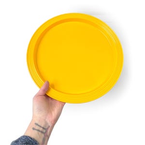 May include: A bright yellow, round serving tray with a ridged edge. The tray is held by a hand against a white background. The tray is a solid, vibrant yellow color.