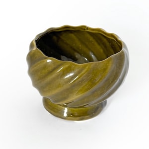May include: A vintage, olive-green ceramic bowl with a pedestal base. The bowl has a spiral design around the exterior and a glossy finish. The interior is dark, and the bowl is set against a white background.