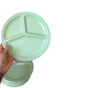May include: A stack of light green divided plates. The plates are round with three compartments. One plate is being held up, showing the design. The plates are made of a durable material and are suitable for everyday use.