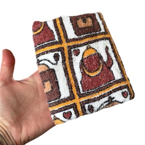 May include: A rectangular, patterned towel with a retro design. The towel features a grid of squares in white, brown, yellow, and maroon. Some squares contain stylized figures and hearts. The towel is held in a hand against a white background.