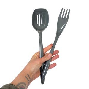 May include: A gray slotted spoon and serving fork set. The spoon has a rectangular head with vertical slots. The fork has four tines. Both utensils have long, slender handles. The set is held against a white background.