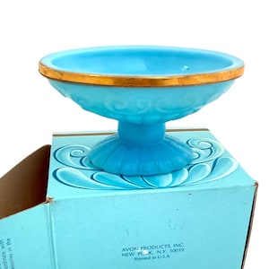 May include: A light blue, pedestal-style dish with a gold rim. The dish sits atop a box with a light blue and white design. The box has text that reads "AVON PRODUCTS, INC. NEW YORK, N.Y. 10019 Printed in U.S.A."