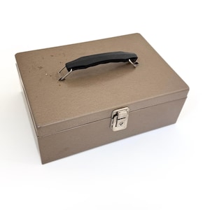 May include: A rectangular, metallic brown storage box with a black handle and a silver latch. The box is closed and sits on a white surface. The handle is centered on the top of the box.