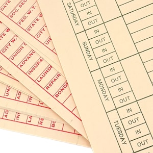 May include: Close-up of vintage time cards with red and black text. The cards are beige and feature sections for recording work hours, including "IN" and "OUT" times for each day of the week. The cards also include sections for deductions.