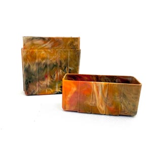 May include: Two rectangular containers with a marbled pattern in shades of orange, brown, and green. One container is taller and divided into sections, while the other is a shorter, open box. Both are set against a white background.