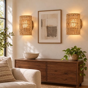 May include: Two cylindrical wall sconces featuring a woven natural fiber shade, each emitting a warm glow. The fixtures are mounted on a wall flanking a framed abstract print above a dark wood console table with a bowl and a potted plant.