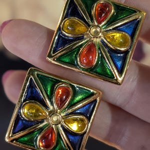 Puede incluir: Un par de pendientes cuadrados con un marco de metal dorado y diseño geométrico. Cada pendiente contiene cuatro cabujones en forma de lágrima de color rojo y amarillo, sobre un fondo de esmalte azul y verde.