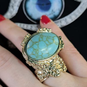 May include: A gold-toned ring featuring a large, oval-shaped turquoise stone with prominent tan veining. The metal band has a textured, organic, and uneven surface finish that surrounds the central stone setting in a decorative manner.