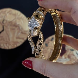 Puede incluir: Un brazalete rígido articulado en tono dorado con un diseño de cabeza de pantera. La cabeza de la pantera está incrustada con pedrería transparente y acentuada con manchas de esmalte negro. La banda de metal dorado texturizado se curva.