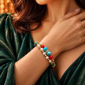 Könnte beinhalten: Ein Perlenarmband mit einer Mischung aus runden türkis-farbenen Steinen, weißen Perlen, facettierten roten Perlen und kleinen metallischen goldfarbenen Zwischenelementen. Das Armband enthält ein Mittelstück mit klaren Kristallen.