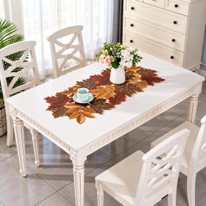 May include: A table runner featuring a collection of fabric autumn leaves in shades of red, orange, brown, and gold. The runner is placed on a white table, accompanied by a white vase with flowers and a small light blue cup on a saucer.