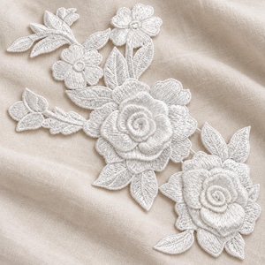 May include: A white floral embroidery patch featuring two large roses and a smaller flower bud, surrounded by detailed leaves and stems. The design is crafted with dense white thread, creating a textured, raised appearance on the fabric surface.