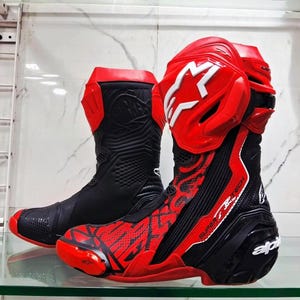 Puede incluir: Un par de botas de moto Alpinestars Supertech en negro y rojo con diseño de corte alto, paneles protectores de plástico moldeado, un logotipo de estrella blanca en la espinillera y detalles perforados en la puntera y los laterales del calzado.
