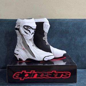 Puede incluir: Un par de botas de moto Alpinestars Supertech R en blanco y negro con construcción de cuero perforado, placas protectoras en las espinillas y sistemas de soporte para el tobillo, colocadas sobre una caja negra de la marca Alpinestars.