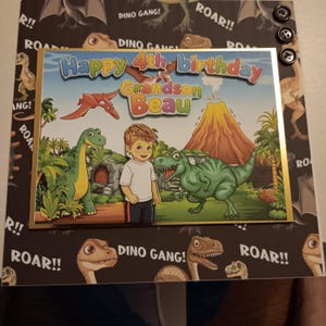 May include: A birthday card featuring a cartoon illustration of a child standing between a green long-necked dinosaur and a green T-Rex in a prehistoric landscape with a volcano. The text reads Happy 4th birthday Grandson Beau.