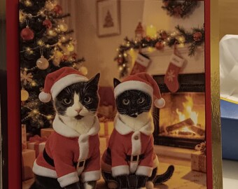 May include: A handmade Christmas greeting card featuring two black and white cats wearing red and white Santa suits and hats. The card has a red border, gold accents, and the text Merry Christmas at the top, with a fireplace and tree background.