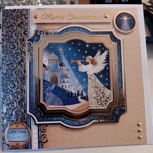 May include: A square greeting card featuring a central layered illustration of an angel playing a trumpet before a blue domed city and a nativity scene under a star. The card includes the text Merry Christmas and Christmas Blessings.