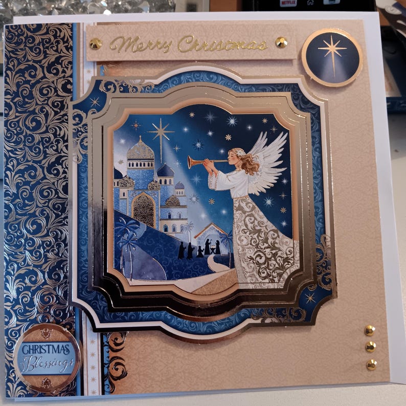May include: A square greeting card featuring a central layered illustration of an angel playing a trumpet before a blue domed city and a nativity scene under a star. The card includes the text Merry Christmas and Christmas Blessings.