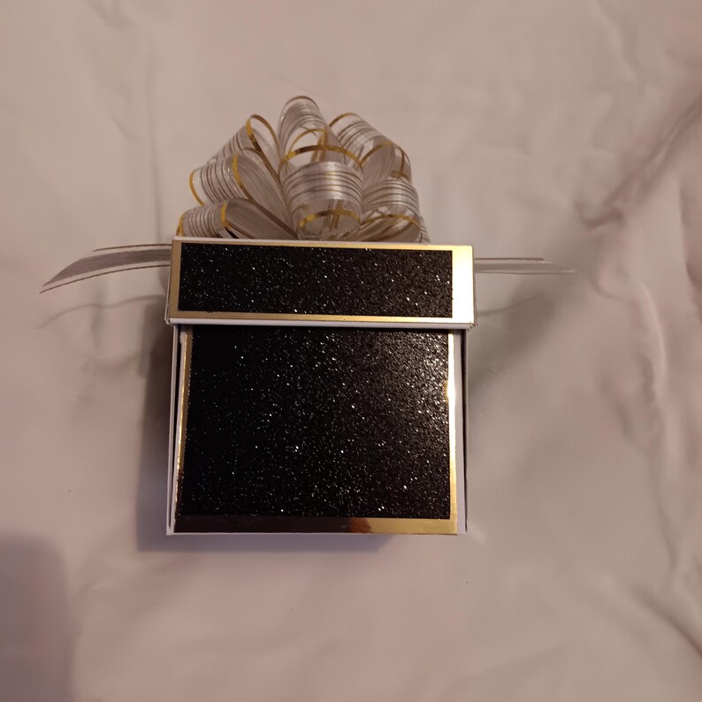 May include: A square gift box featuring a black glittery finish on the lid and sides, accented with gold metallic trim. A large, multi-looped decorative bow made of translucent white ribbon with thin gold stripes sits on top of the box.
