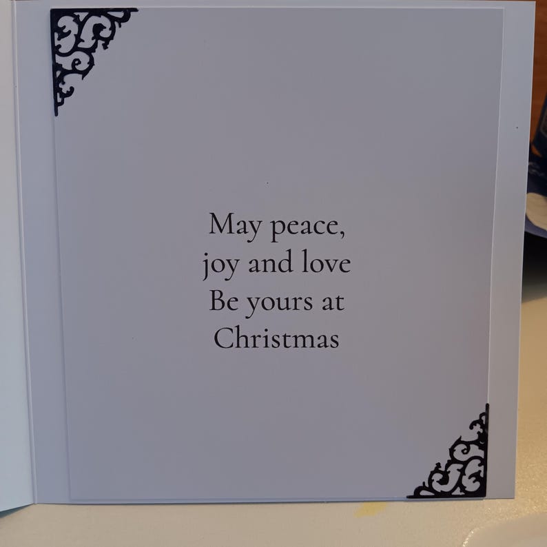 May include: A white greeting card with black filigree corner accents. The center text reads: May peace, joy and love Be yours at Christmas. The card features a clean, simple layout with dark decorative flourishes in the top left and bottom right.