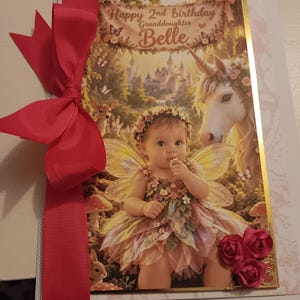 May include: A rectangular greeting card featuring a fairy-themed illustration of a child with wings, a unicorn, and a deer. The text reads Happy 2nd birthday Granddaughter Belle. A large red ribbon is attached to the left side, and three red paper roses are at the bottom right corner.