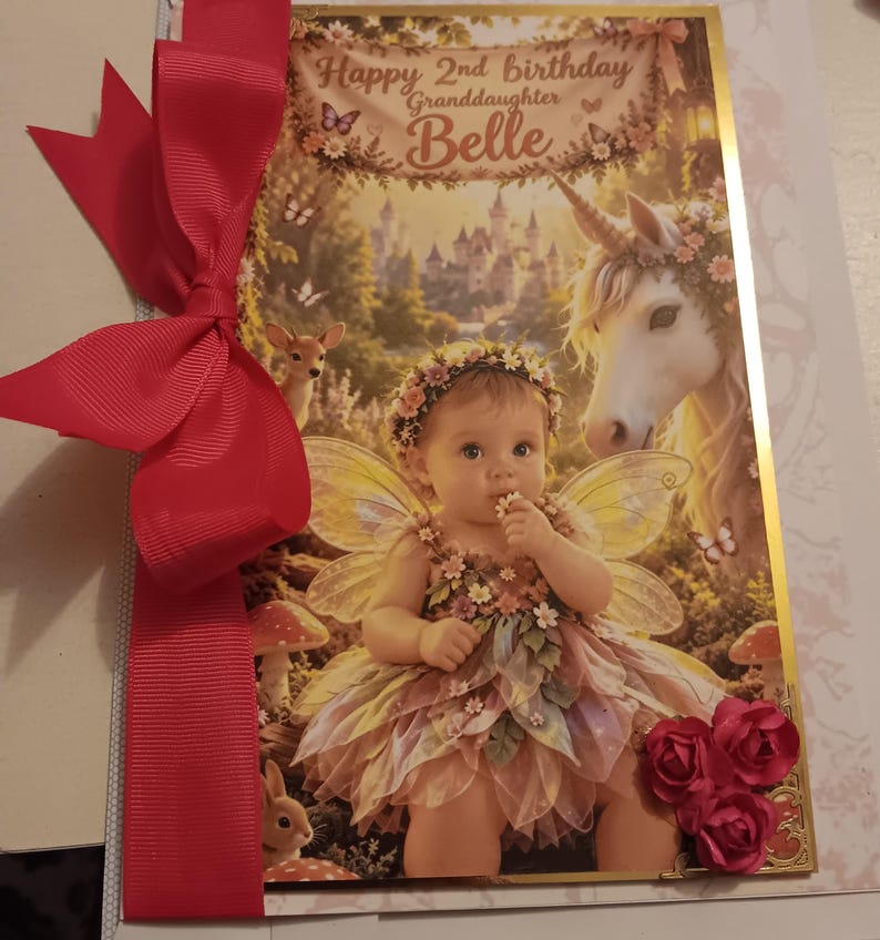 May include: A rectangular greeting card featuring a fairy-themed illustration of a child with wings, a unicorn, and a deer. The text reads Happy 2nd birthday Granddaughter Belle. A large red ribbon is attached to the left side, and three red paper roses are at the bottom right corner.