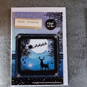 May include: A rectangular Christmas greeting card featuring a blue and purple snowy night scene. A black frame highlights a silhouette of a deer and Santa in a sleigh with reindeer against a full moon. Text reads Happy Christmas and Santa is on his way.
