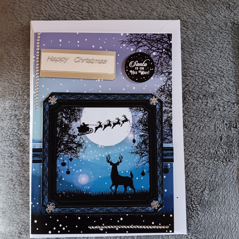 May include: A rectangular Christmas greeting card featuring a blue and purple snowy night scene. A black frame highlights a silhouette of a deer and Santa in a sleigh with reindeer against a full moon. Text reads Happy Christmas and Santa is on his way.