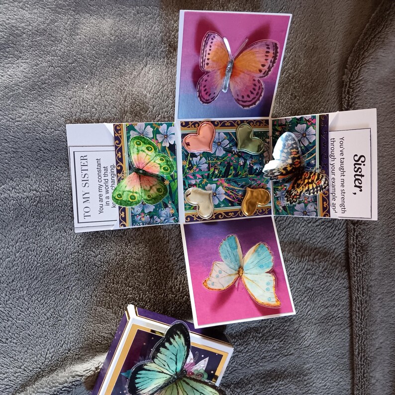 May include: A square gift box that unfolds into a cross shape, featuring colorful butterfly cutouts, metallic heart charms, and printed text. The panels display floral patterns and messages including 'TO MY SISTER' and 'Sister, you've taught me strength'.