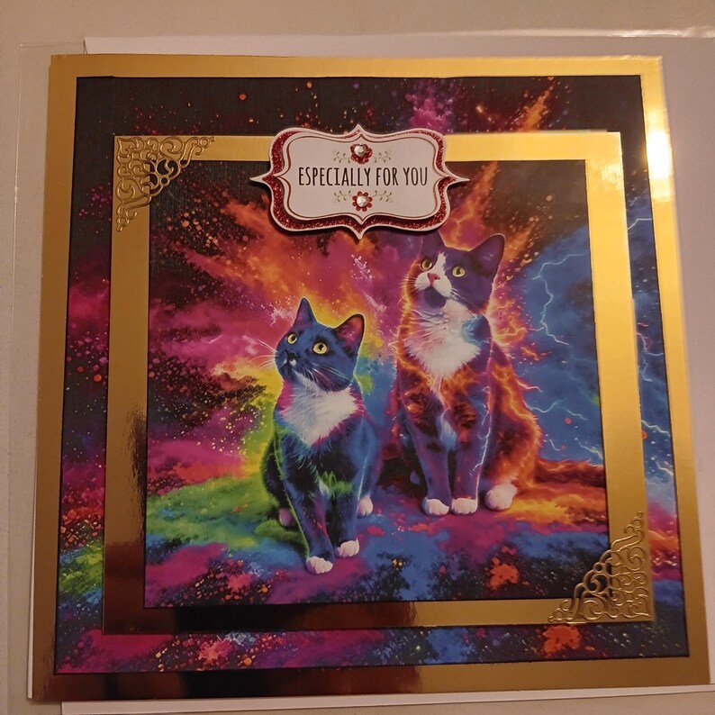 May include: A square greeting card featuring two cats against a vibrant, multicolored cosmic background with lightning effects. The card is framed in gold with decorative corner accents and a central label that reads ESPECIALLY FOR YOU.