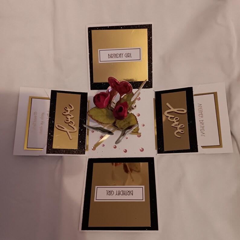 May include: A square explosion greeting card featuring black, white, and gold panels. The center displays red fabric roses and a heart. Text panels read Birthday Girl, Love, and Another Birthday in black lettering on white and gold backgrounds.