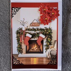 May include: A rectangular Christmas greeting card featuring a fireplace scene with a green garland, two red stockings labeled Mary and Tammy, and a sign reading Merry Christmas. Three red poinsettia flowers are attached to the top right corner.