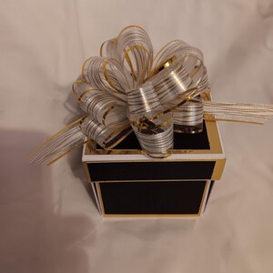 May include: A square gift box with a matte black finish and gold metallic trim. The lid is topped with a large, multi-looped decorative bow made of sheer white ribbon featuring thin, horizontal gold metallic stripes and gold-edged borders.