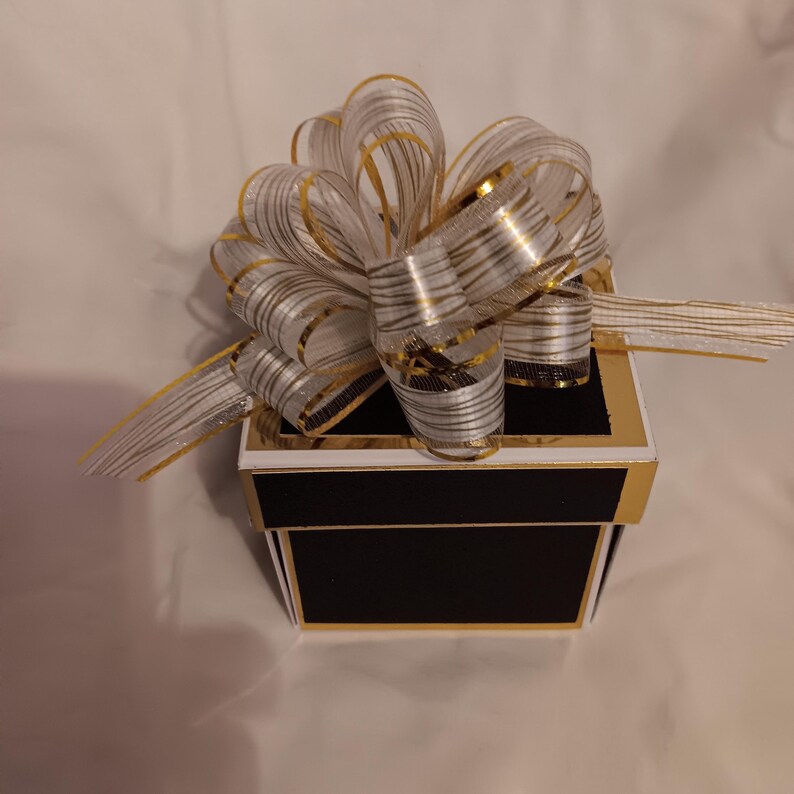 May include: A square gift box with a matte black finish and gold metallic trim. The lid is topped with a large, multi-looped decorative bow made of sheer white ribbon featuring thin, horizontal gold metallic stripes and gold-edged borders.