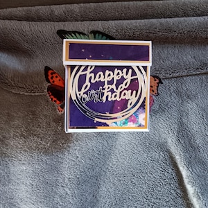 May include: A square gift box featuring a deep purple lid and a front panel decorated with a gold circular cutout that displays the text Happy Birthday in a cursive script, accented by two butterfly wings extending from the sides.