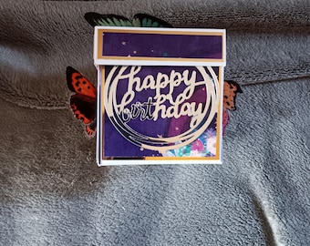 May include: A square gift box featuring a deep purple lid and a front panel decorated with a gold circular cutout that displays the text Happy Birthday in a cursive script, accented by two butterfly wings extending from the sides.