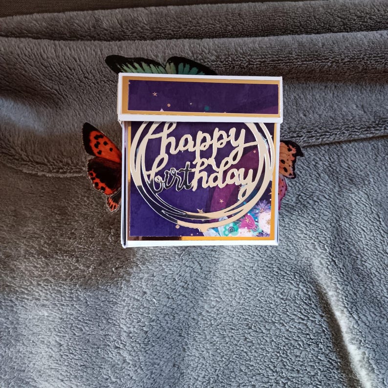 May include: A square gift box featuring a deep purple lid and a front panel decorated with a gold circular cutout that displays the text Happy Birthday in a cursive script, accented by two butterfly wings extending from the sides.