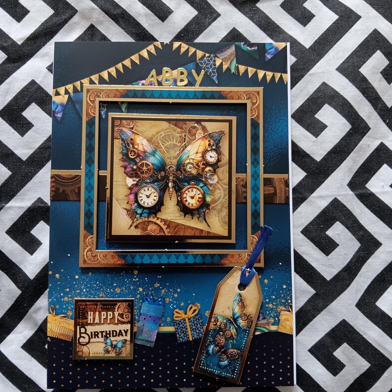 May include: A rectangular birthday card featuring a central square frame with a mechanical butterfly illustration, clock faces, and gears. The card displays the name ABBY, gold bunting, gift boxes, and a hanging tag with a butterfly design.