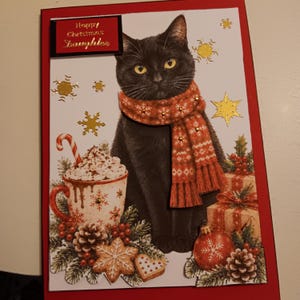 May include: A rectangular Christmas greeting card featuring a black cat wearing a red patterned scarf. The card displays the text Happy Christmas Daughter, a mug of hot cocoa with a candy cane, gold foil snowflakes, pinecones, and gifts.