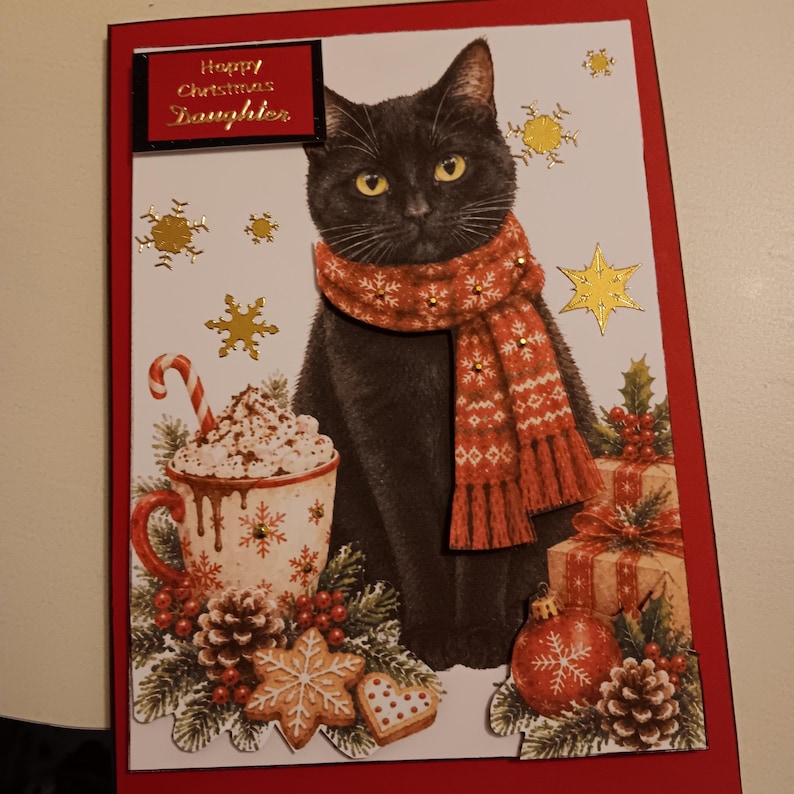 May include: A rectangular Christmas greeting card featuring a black cat wearing a red patterned scarf. The card displays the text Happy Christmas Daughter, a mug of hot cocoa with a candy cane, gold foil snowflakes, pinecones, and gifts.