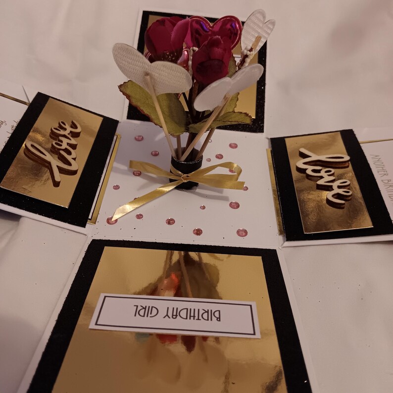 May include: A pop-up greeting card featuring a central bouquet of red fabric roses and white heart-shaped cutouts. The card has gold metallic panels with the word Love in wooden script, and a white panel with the text BIRTHDAY GIRL.