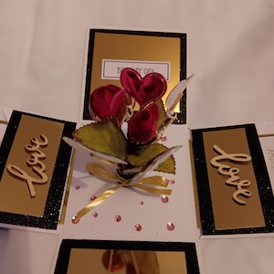 May include: A square pop-up greeting card featuring a central bouquet of red roses and a shiny pink heart. The card has black and gold panels, with the word "love" in wooden script on two sides and "BIRTHDAY GIRL" text at the top.