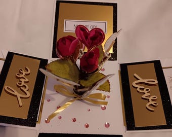 May include: A square pop-up greeting card featuring a central bouquet of red roses and a shiny pink heart. The card has black and gold panels, with the word "love" in wooden script on two sides and "BIRTHDAY GIRL" text at the top.