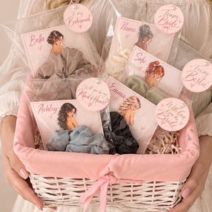 May include: A white woven basket with a pink fabric liner contains several hair scrunchies in clear plastic packaging. Each package features a card with an illustration of a person and text including names like Bella, Sofi, Ashley, and Emma, plus phrases like To my Bridesmaid, Team Bride, and Will you be my Maid of Honor?