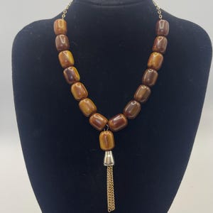 Könnte beinhalten: Eine Halskette mit glatten, zylindrischen braunen Perlen in Marmoroptik, die auf einer goldfarbenen Kette aufgereiht sind. In der Mitte befindet sich eine einzelne Perle an einer silberfarbenen Metallkappe mit einer goldenen Kettenquaste.