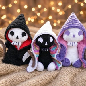 May include: Three crocheted figures with hoods and scythes. The left figure is black with a red inner hood. The centre figure is black with a white hood featuring rainbow-coloured lining. The right figure is white with a purple hood and pink lining.