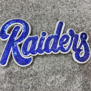 May include: A decorative patch featuring the word Raiders written in a cursive script. The lettering is filled with small, reflective blue sequins and outlined with a thick, white border that follows the shape of the text.