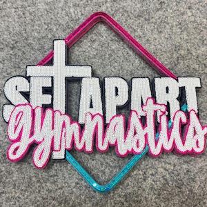 May include: A layered sign featuring a diamond-shaped frame with a pink top and blue bottom. The white text reads SET APART with a cross replacing the letter T, and the word gymnastics is written in a pink cursive script font below.