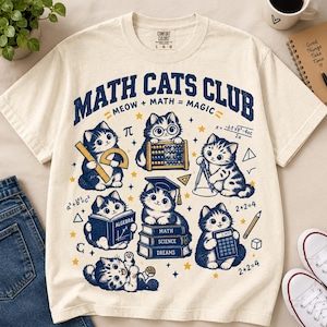 May include: A cream-colored short-sleeve t-shirt featuring a dark blue graphic print with the text MATH CATS CLUB and MEOW + MATH = MAGIC. The design includes six illustrated cats with math tools, books, equations, and geometric shapes.